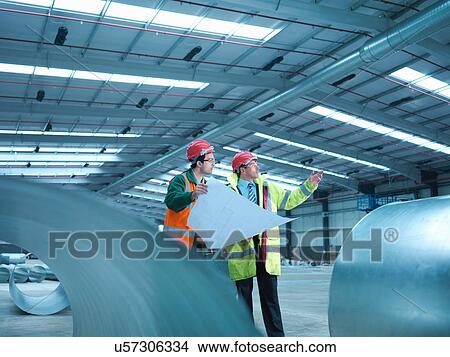 Picture - Site foreman advising apprentice with plans in partly completed factory. Fotosearch