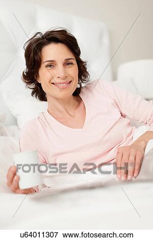 Stock Photo - Smiling woman reading newspaper in bed. Fotosearch