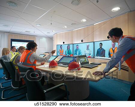 Stock Image - Staff members in meeting room with colleagues on video conference screens. Fotosearch