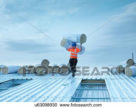 Stock Image - Workman carrying rolls of insulation to install into factory roof. Fotosearch