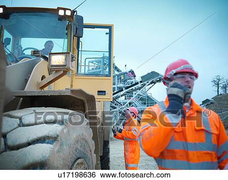 Stock Photograph - Workman using walkie talkie next to industrial digger on quarry site. Fotosearch