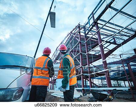 Workmen watching crane lifting roof panels onto steel construction frame on building site View Large Photo Image Stock Photo - Workmen watching crane lifting roof panels onto steel construction frame on building site. Fotosearch