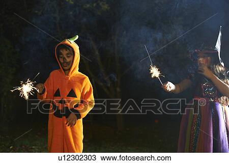 Stock Image - Brother and sister wearing halloween costumes holding sparklers in garden at night. Fotosearch