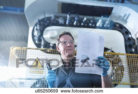 Stock Photo - Engineer holding plans in front of CNC machine in factory. Fotosearch