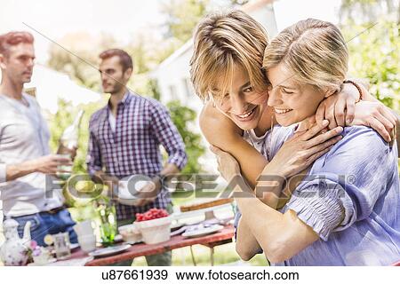 Stock Photo - "Group of friends setting up garden party, two female friends embracing". Fotosearch