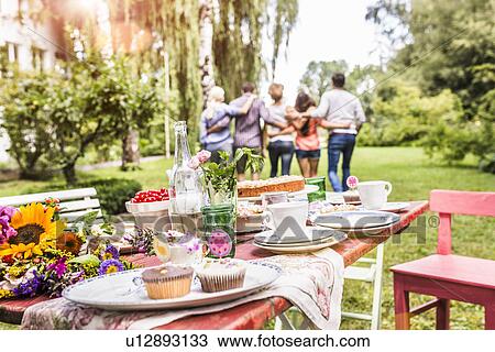 Stock Image - "Group of friends walking away with arms around each other, garden party table with food in foreground". Fotosearch
