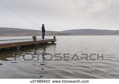 Stock Image - "Man standing alone on pier looking out to sea, Vifilsstadavatn, Iceland". Fotosearch