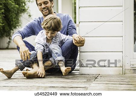 Stock Photo - Mid adult man and son laughing and tickling feet on porch. Fotosearch