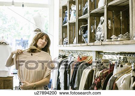 Stock Image - Mid adult woman holding dress in front of herself in vintage boutique. Fotosearch