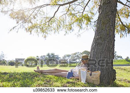 Stock Image - "Mid adult woman sitting against tree, sleeping". Fotosearch