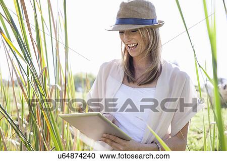 Stock Image - "Mid adult woman sitting in long grass, using digital tablet". Fotosearch