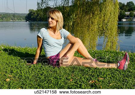 Stock Photo - Mid adult woman stretching on grass beside lake. Fotosearch