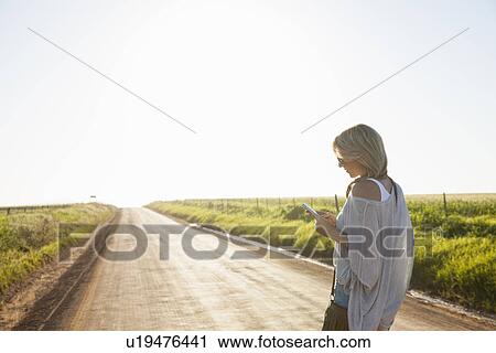 "Mid adult woman walking on country road, looking at mobile phone" View Large Photo Image Stock Image - "Mid adult woman walking on country road, looking at mobile phone". Fotosearch