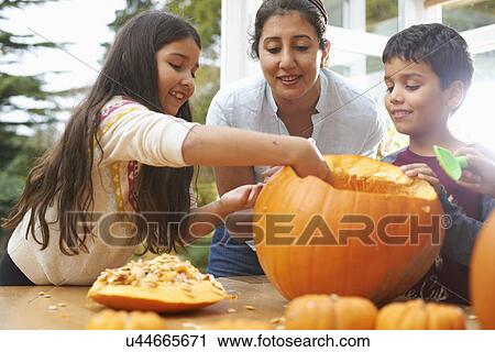 Stock Image - Mother and children hollowing out pumpkin in dining room. Fotosearch