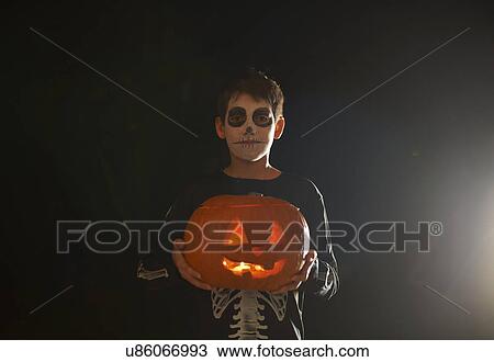 Stock Image - Portrait of boy wearing halloween skeleton costume holding pumpkin. Fotosearch