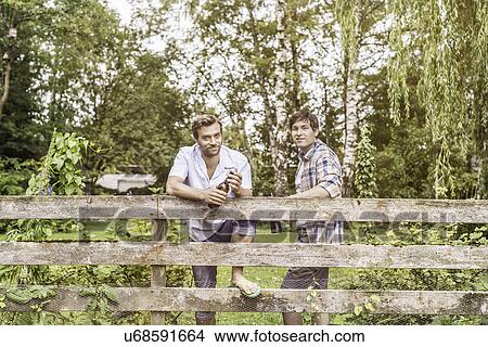 Picture - Portrait of two male adult friends drinking beer at garden fence. Fotosearch