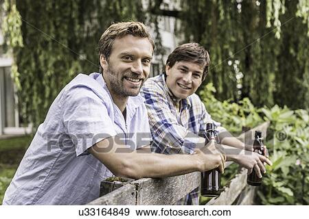 Stock Photo - Portrait of two male adult friends leaning on garden fence drinking beer. Fotosearch