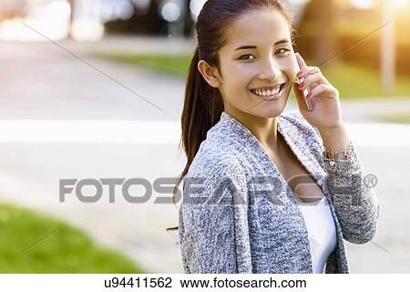 Stock Image - Portrait of young woman chatting on smartphone in park. Fotosearch