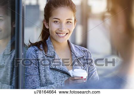 Picture - Portrait of young woman drinking takeaway coffee leaning against park building. Fotosearch