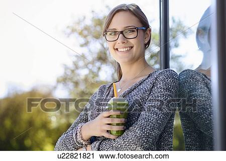 Stock Image - Portrait of young woman with takeaway coffee leaning against park building. Fotosearch