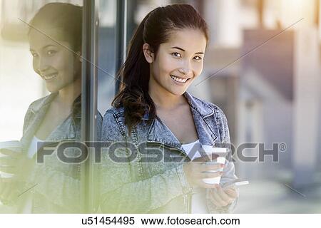 Stock Photography - Portrait of young woman with takeaway coffee and smartphone leaning against park building. Fotosearch