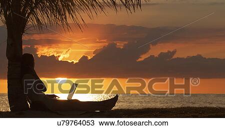 Stock Image - "Silhouette of young woman with laptop looking at sunset over sea, Etosha National Park, Namibia". Fotosearch