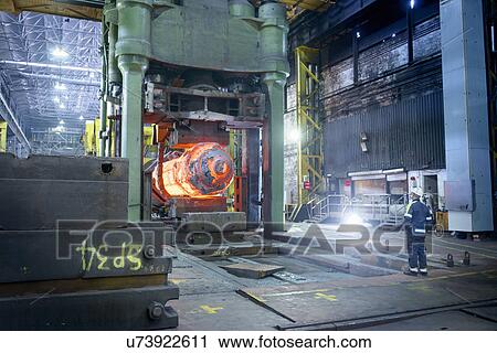 Stock Image - Steelworker inspects hot steel in forging press in steelworks. Fotosearch