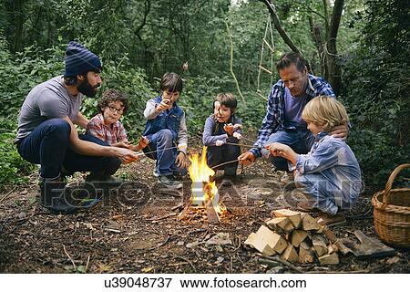 Stock Photo - Two fathers and four boys toasting marshmallows on campfire in forest. Fotosearch