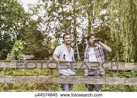 Stock Photography - Two male adult friends drinking beer at garden fence. Fotosearch