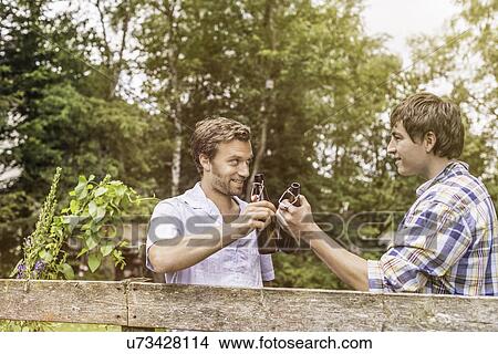 Picture - Two male adult friends toasting with beer bottles at garden fence. Fotosearch