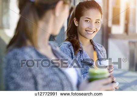 Stock Image - Two young women with takeaway coffee leaning against park building. Fotosearch