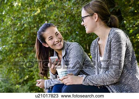 Stock Image - Two young women with takeaway coffee sitting chatting in park. Fotosearch