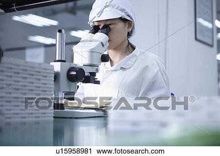 Stock Image - Worker using microscope in factory that specialises in creating functional circuits on flexible surfaces. Fotosearch