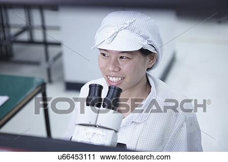 Stock Image - Worker using microscope in factory that specialises in creating functional circuits on flexible surfaces. Fotosearch