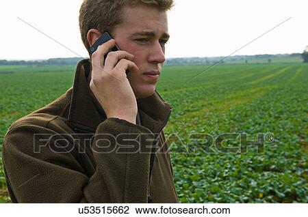 Stock Image - Young male farmer chatting on smartphone in field. Fotosearch