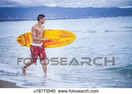 Stock Image - "Young male surfer running with surfboard on beach, Cagliari, Sardinia, Italy". Fotosearch