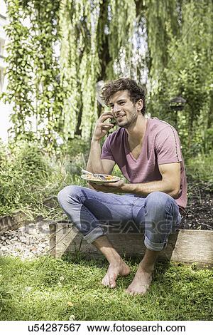 Young man in garden on cell phone with plate of food View Large Photo Image Stock Photo - Young man in garden on cell phone with plate of food. Fotosearch