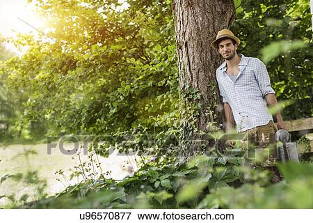 Young man leaning against tree with watering can View Large Photo Image Stock Photo - Young man leaning against tree with watering can. Fotosearch