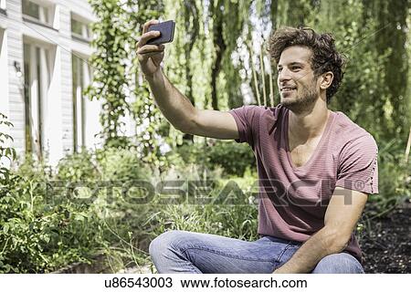 Stock Image - Young man taking selfie with cell phone in garden. Fotosearch