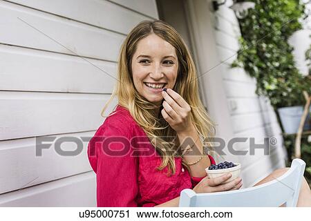 Stock Image - Young woman eating blueberries in front porch. Fotosearch