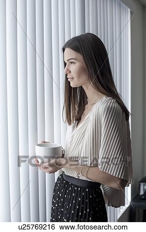 Stock Photograph - Young woman looking out of window with coffee. Fotosearch