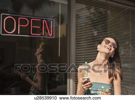 "Young woman standing outside shop, holding smartphone, laughing" View Large Photo Image Stock Image - "Young woman standing outside shop, holding smartphone, laughing". Fotosearch