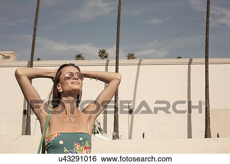 Stock Photograph - "Young woman wearing sunglasses, hands behind head, looking up". Fotosearch