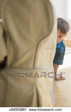 Stock Image - Boy (6-7) peeking around side of sofa. Fotosearch