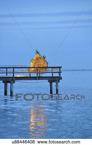 Stock Photo - "Christmas tree with yellow lights on pier, blue sky and water at dusk". Fotosearch