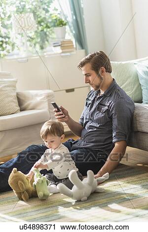 Stock Image - Father sitting on floor with little son (2-3 years) in living room and texting message. Fotosearch