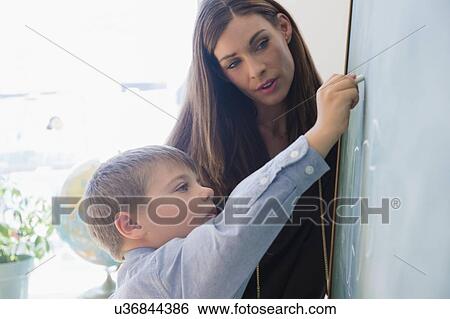 Female teacher assisting schoolboy (6-7) while writing on blackboard View Large Photo Image Stock Photograph - Female teacher assisting schoolboy (6-7) while writing on blackboard. Fotosearch