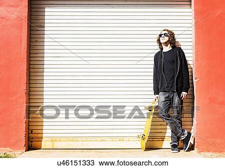 Stock Image - Man with skateboard leaning on garage door. Fotosearch