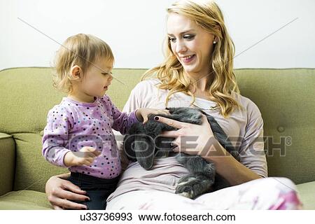 Stock Image - Mother and daughter (12-17 months) sitting on sofa stroking rabbit. Fotosearch