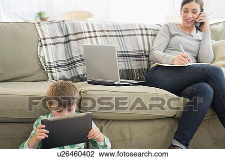 Stock Image - Mother and son (6-7) sitting in living room with tablet and laptop. Fotosearch